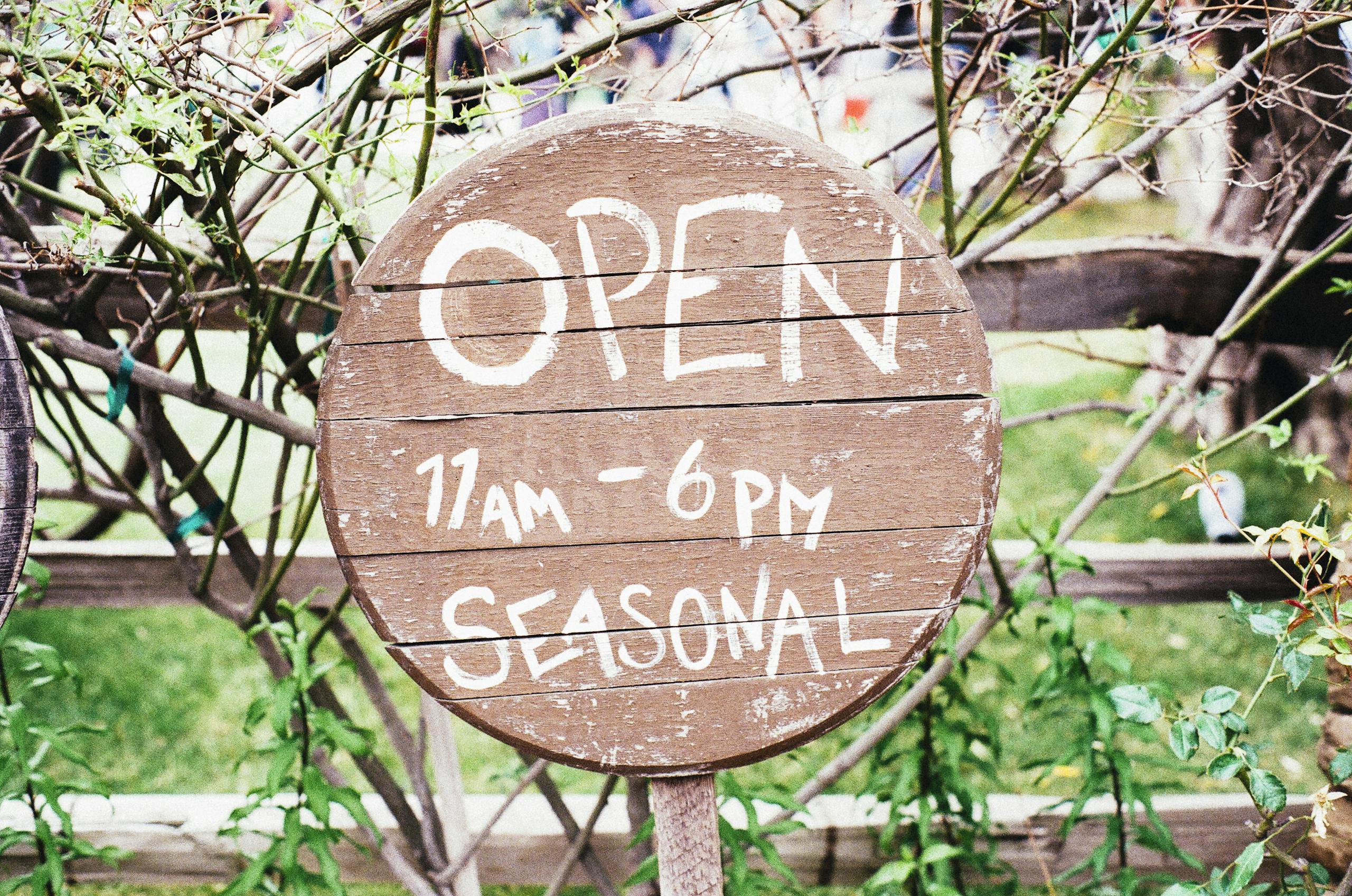 Round wooden sign displaying 'Open 11am-6pm Seasonal' amidst greenery.
