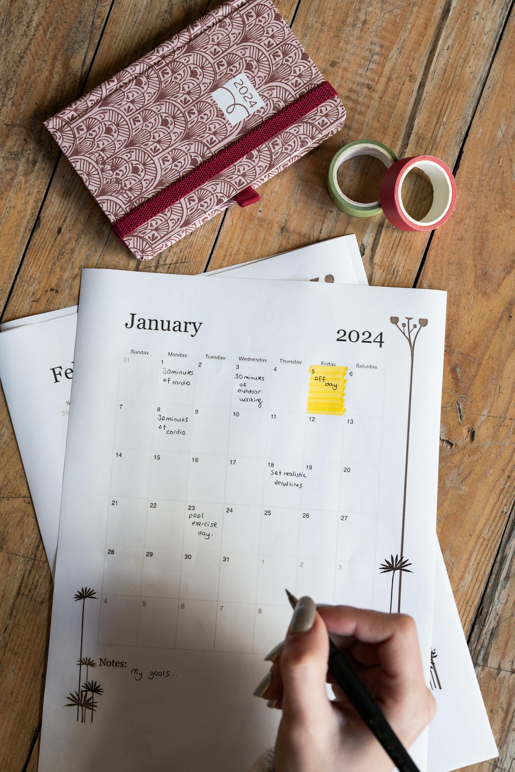 Close-up of a hand writing on a January 2024 calendar page with a notebook on a wooden table.